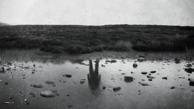 Uma foto em preto e branco de uma margem vazia de um rio, com a figura de um homem refletida na água