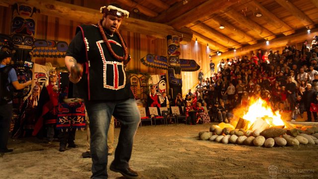 A man wearing a traditional indigenous shirt, hat, and neckpiece dances around a firepit in a large wooden lodge as a crowd of people watch