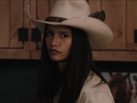 Avery stands in the Yellowstone bunkhouse wearing a white cowboy hat looking confrontational